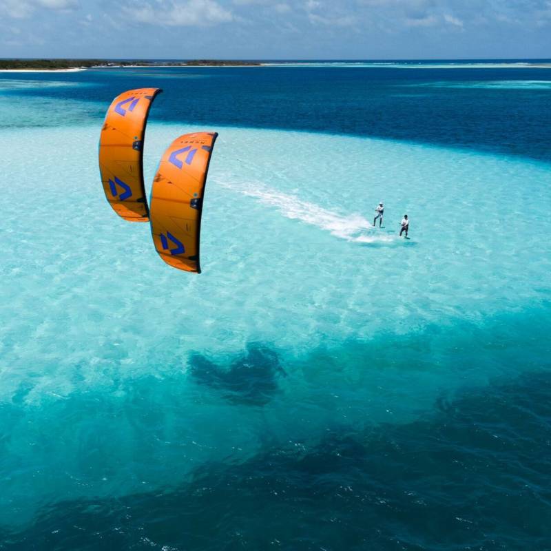 Deux riders pratiquant le kitesurf sur une mer calme, sous une météo ensoleillée, avec une plage en arrière-plan.