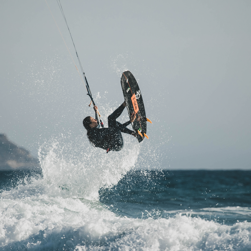 Kitesurfeur effectuant un saut aérien avec une planche F-ONE Magnet Carbon 2023 noire sur une mer agitée, équipé d'une combinaison intégrale