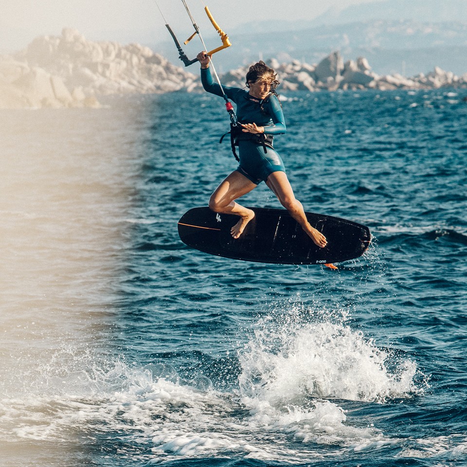 Kitesurfeuse réalisant une figure freestyle avec la planche F-ONE Magnet Carbon 2023 noire, sur une mer calme près d’un littoral rocheux, en combinaison turquoise
