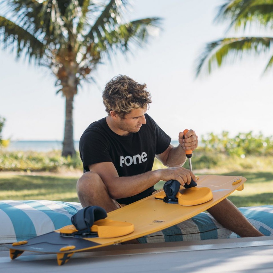 Un kitesurfeur règle ses Pads F-ONE Platinium 3 sur une planche de kitesurf sous des palmiers à Madagascar. Il utilise un tournevis pour ajuster les straps de kitesurf, mettant en avant le système de réglage personnalisable et la facilité d'installation de ces pads bi-densité conçus pour un confort optimal et une connexion parfaite en kitesurf.