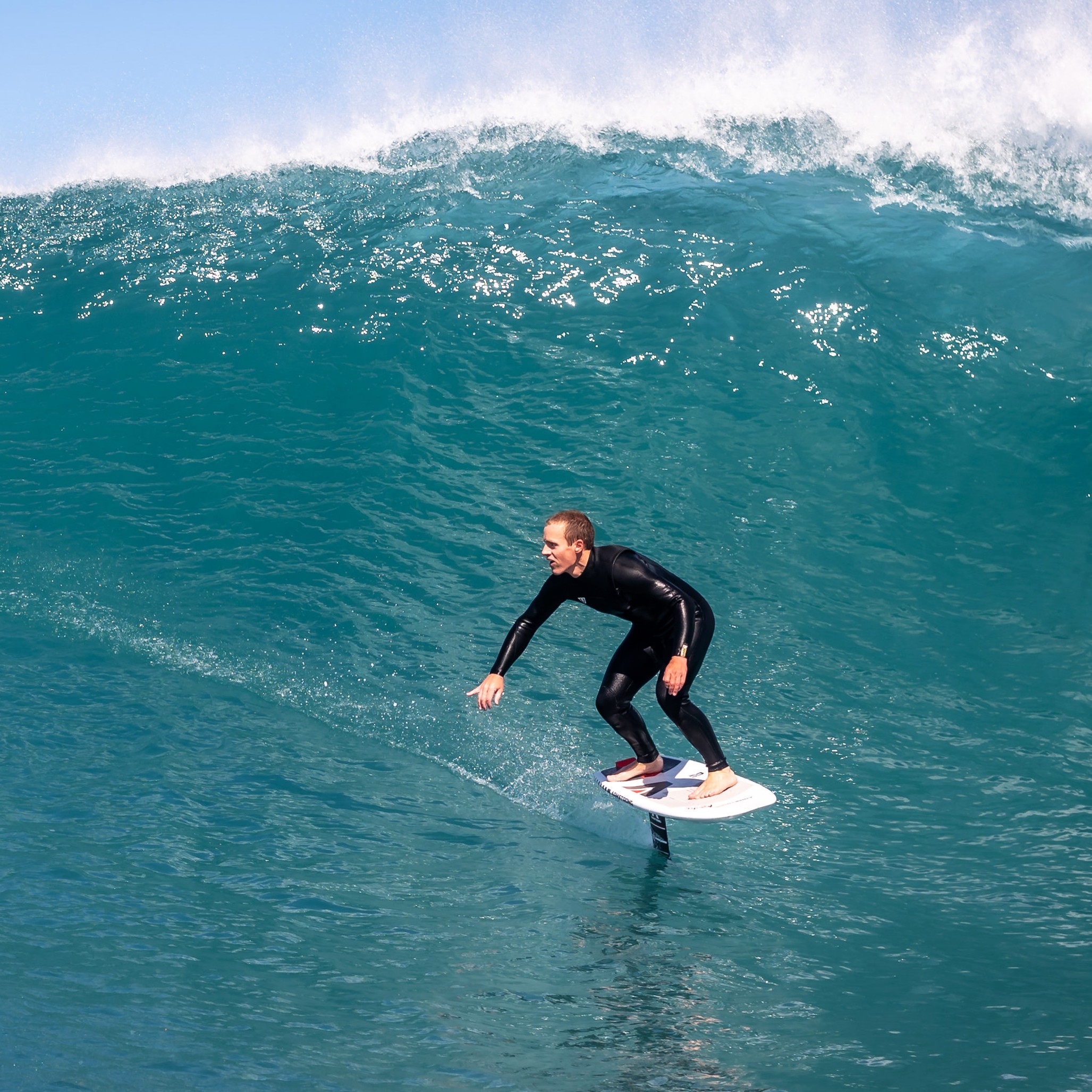 Rider pratiquant le surf foil sur une grande vague avec une planche équipée d'un foil, sur une mer bleue claire sous un ciel dégagé