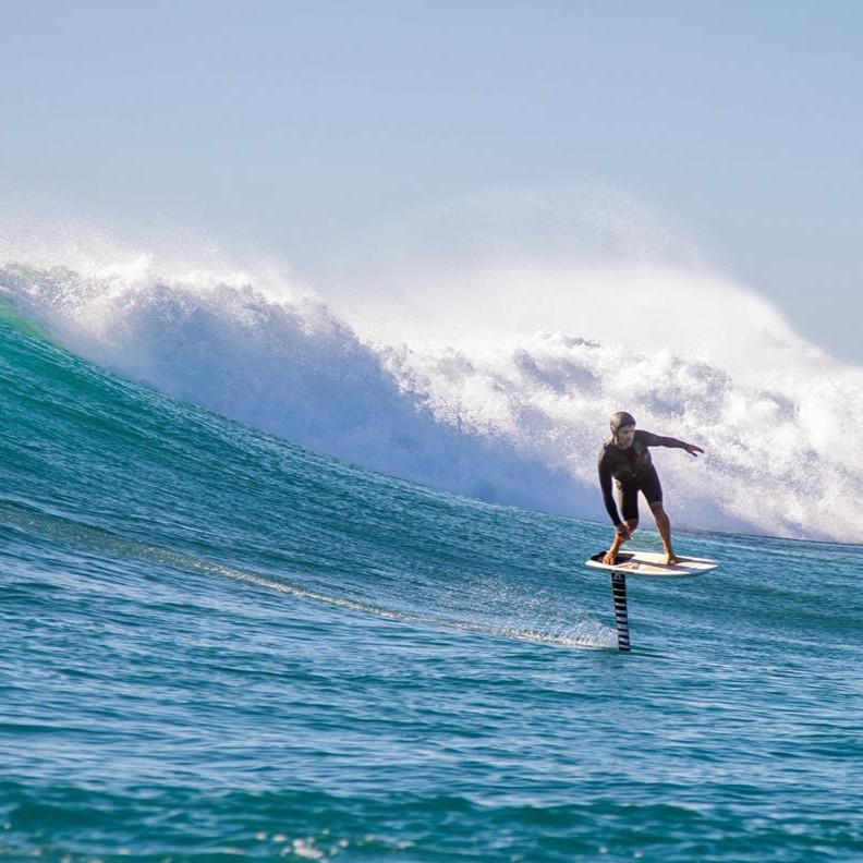 Surfeur en combinaison noire pratiquant le surf foil, chevauchant une planche équipée d'un foil, avec une grande vague en arrière-plan