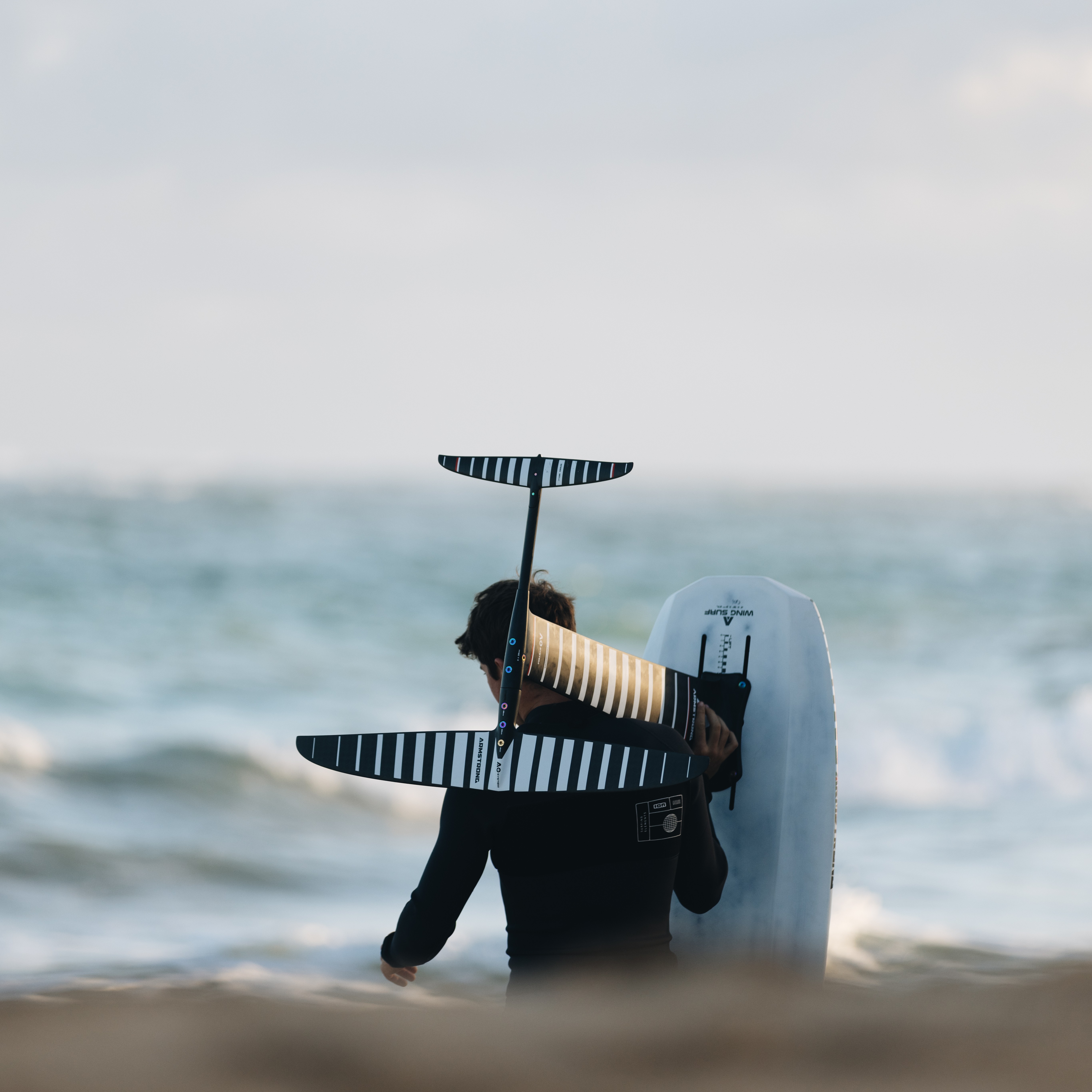 Surfeur portant son équipement de foil, avec le foil et la planche sur l'épaule, marchant sur la plage avec l'océan en arrière-plan