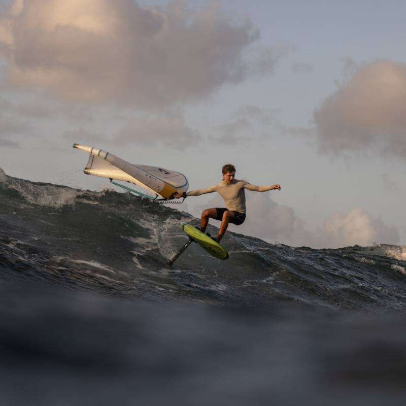 Pratiquant de wingfoil sur planche à foil verte, naviguant sur une grosse vague au coucher du soleil