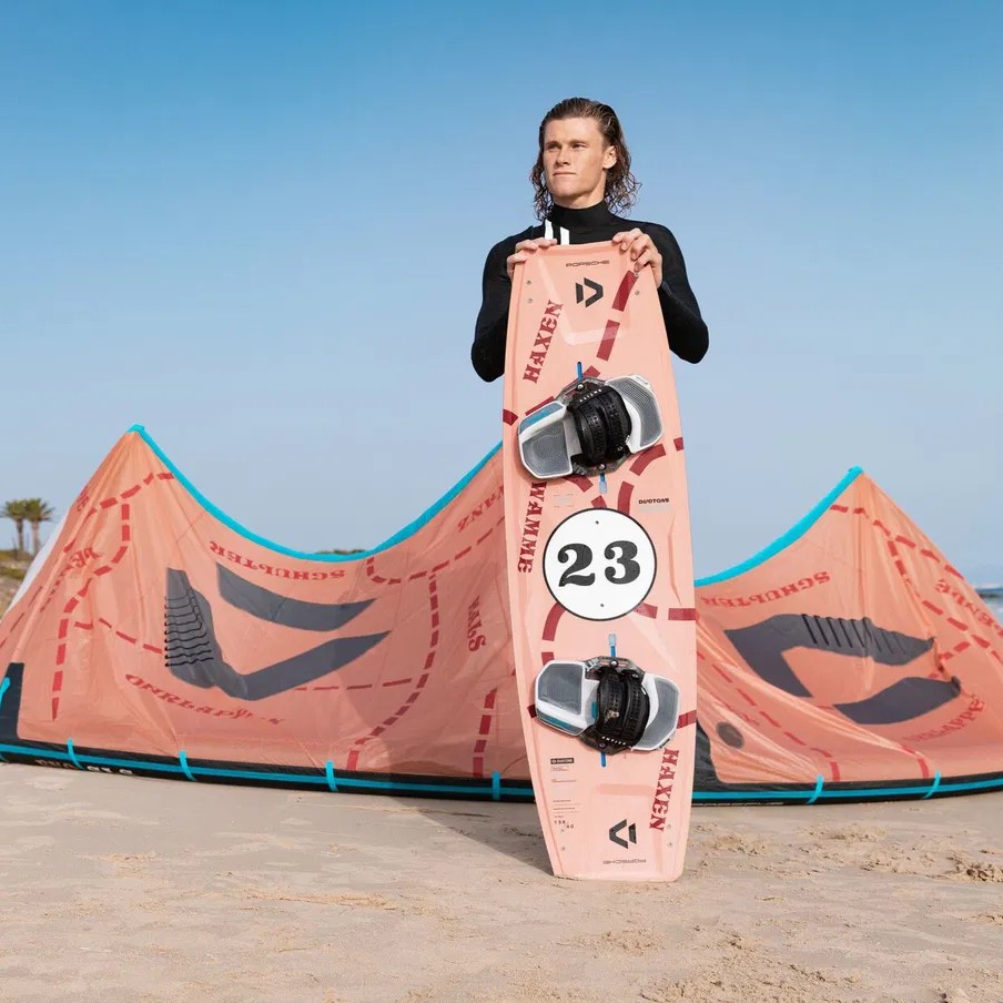Rider présentant la planche de kitesurf Duotone Select Concept Blue Pink Pig Limited Edition 2025 sur la plage devant l’aile de kite assortie