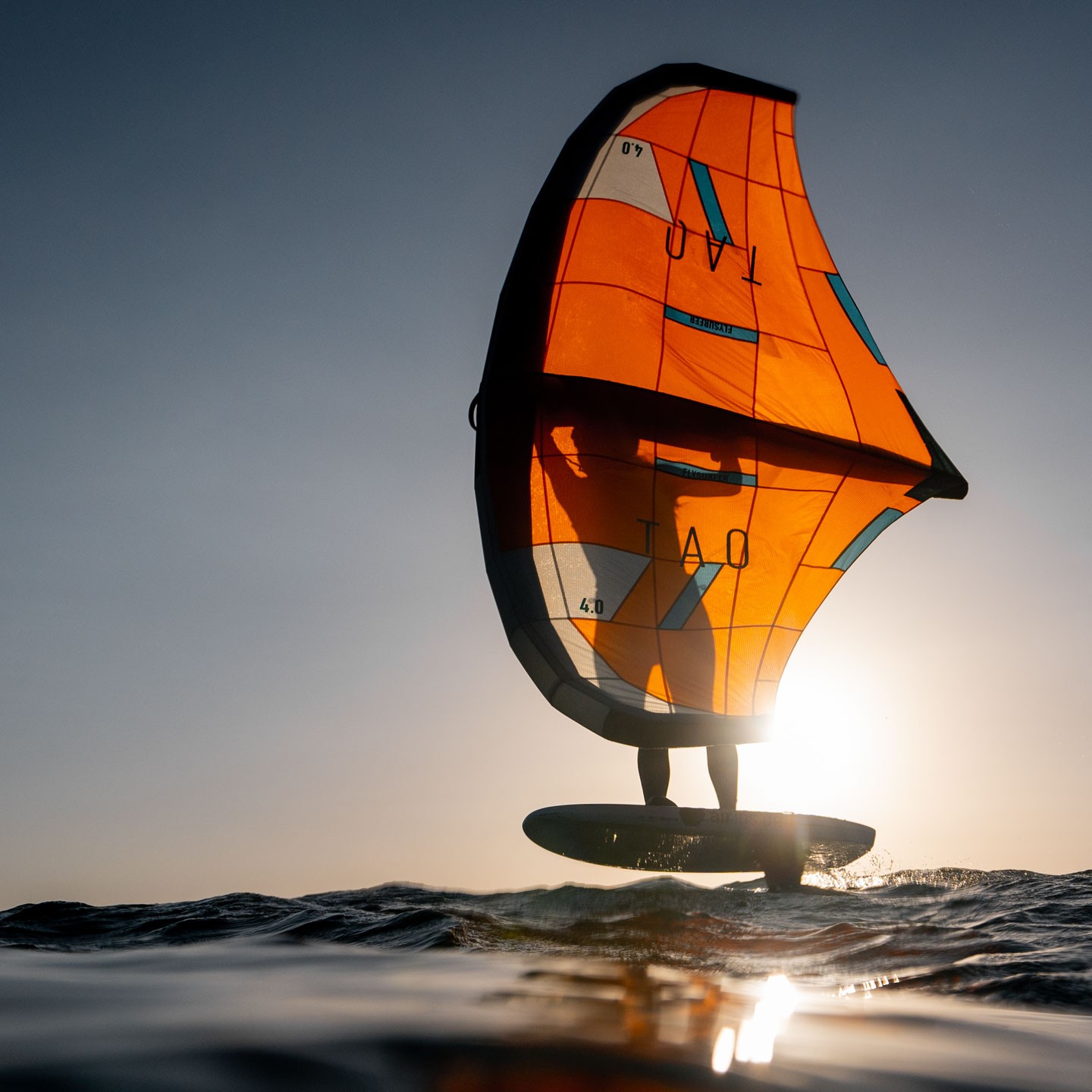 Rider effectuant une figure freestyle en wingfoil avec une aile orange Flysurfer sur fond de ciel dégagé et vague océanique – performance extrême en mer avec équipement de kitesurf 2025.