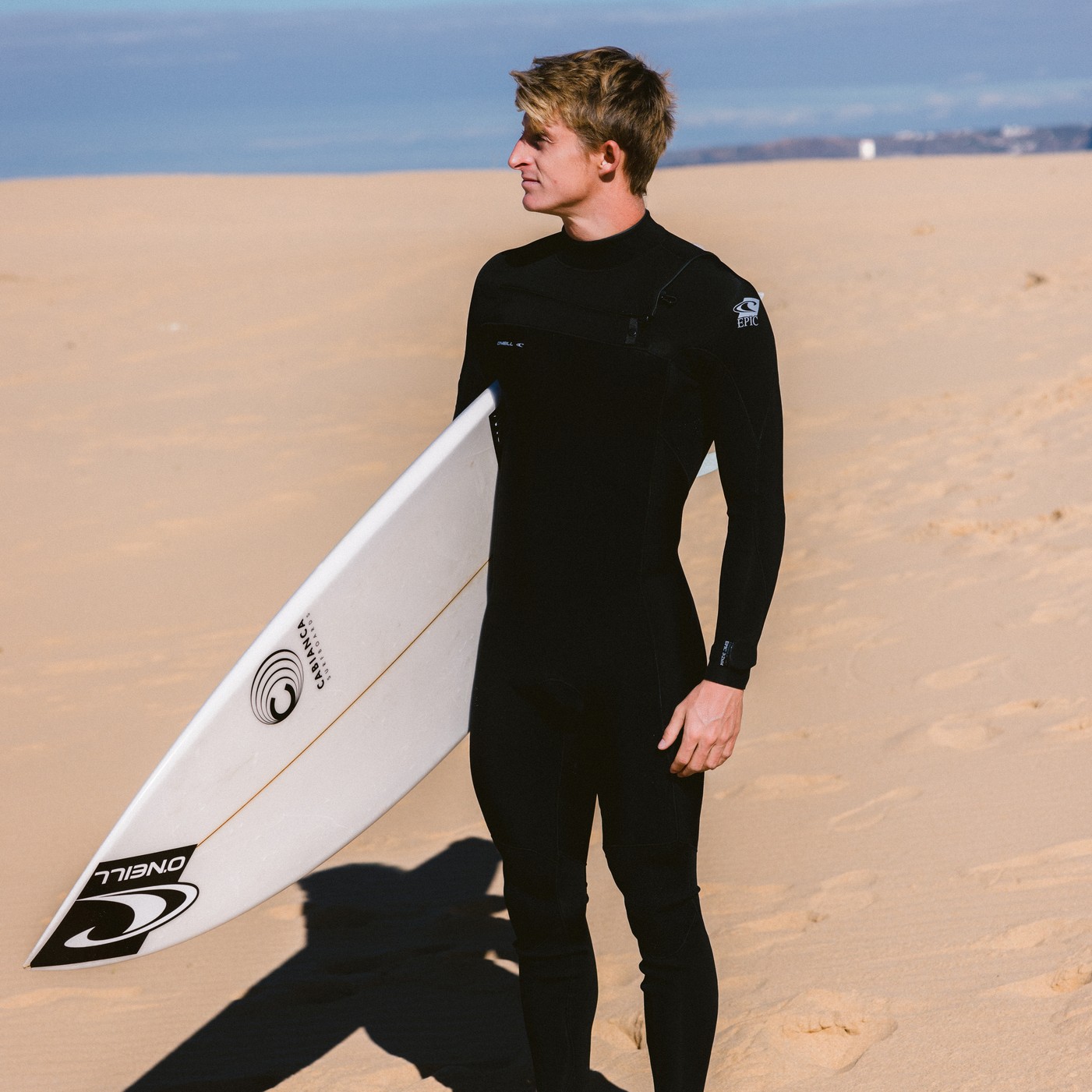 Homme portant la combinaison O'Neill Epic 3/2 Chest Zip Full 2025 sur la plage avec planche de surf à la main