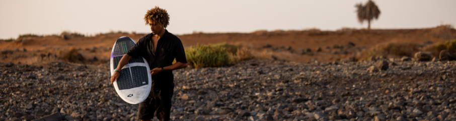 Rider de kitesurf transportant son surf équipé de pads, avant une session strapless sur spot rocheux